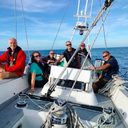 adult crew posing behind helm for group photo, bright blue skies behind
